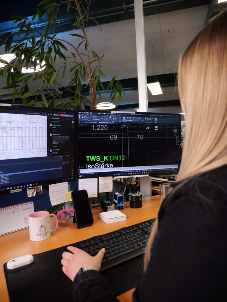 Frau arbeitet an Computer mit mehreren Monitoren im Büro, umgeben von Pflanzen und Büromaterialien.