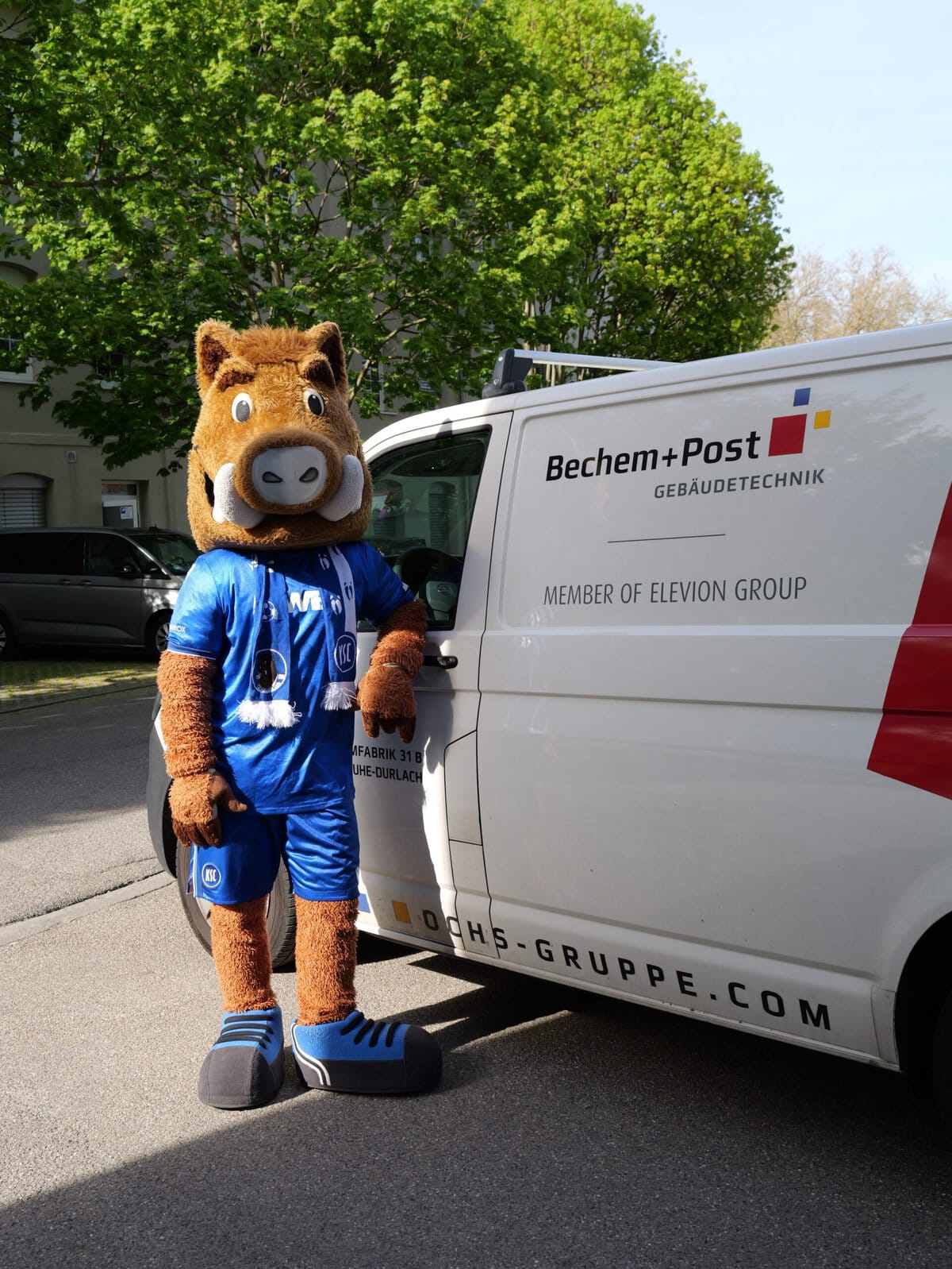 Maskottchen von Bechem + Post neben Firmenfahrzeug auf der Straße.